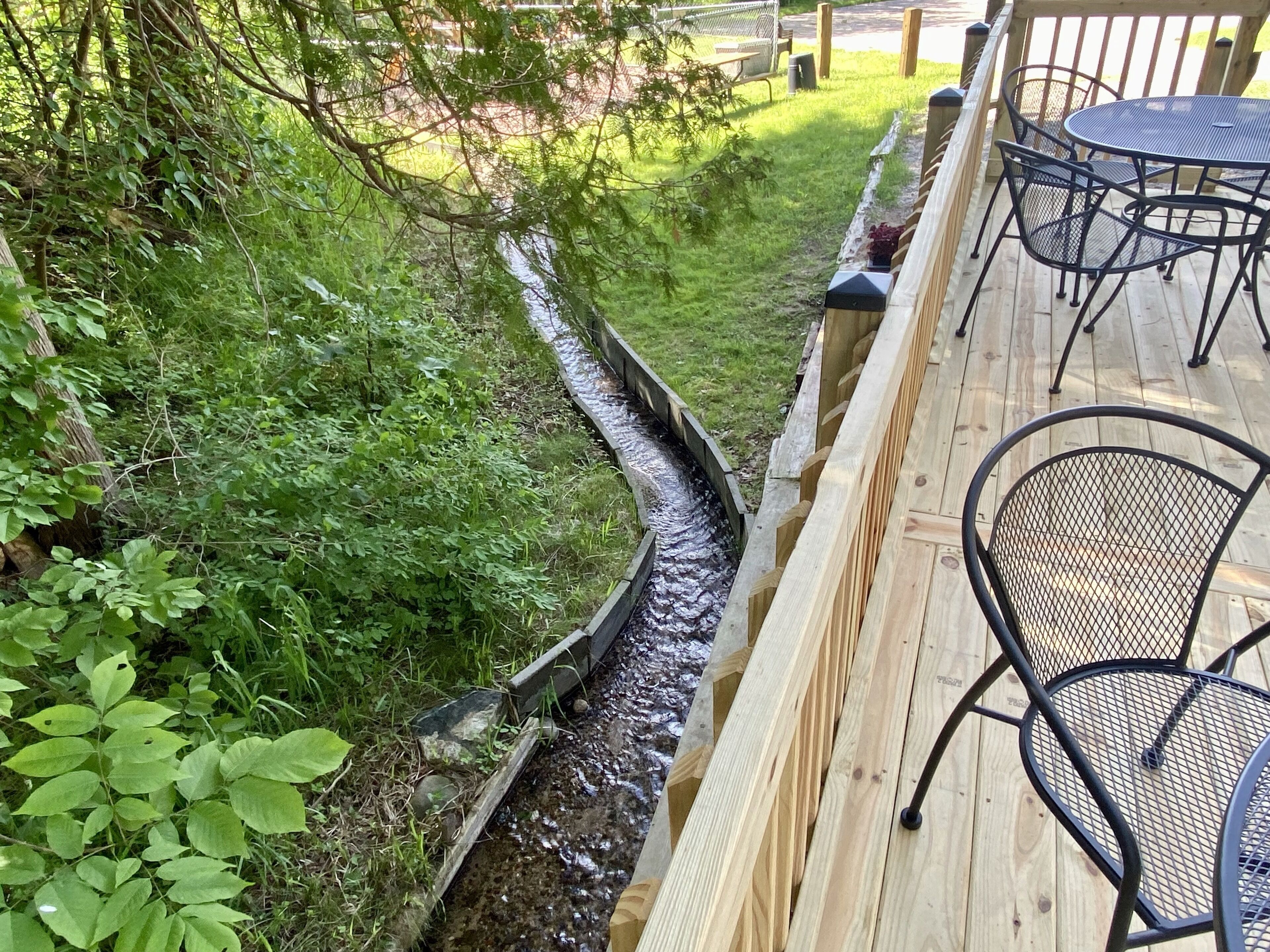 A wooden deck with metal chairs and a table, surrounded by lush greenery and a narrow water channel.