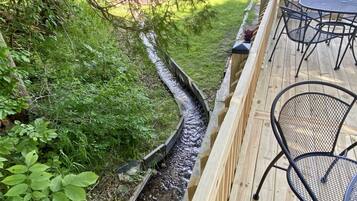 A wooden deck with metal chairs and a table, surrounded by lush greenery and a narrow water channel.