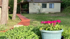 A small house with a red pathway leading to a porch, surrounded by lush greenery and a large flower pot. - Betsie Riverside Resort (Benzonia)