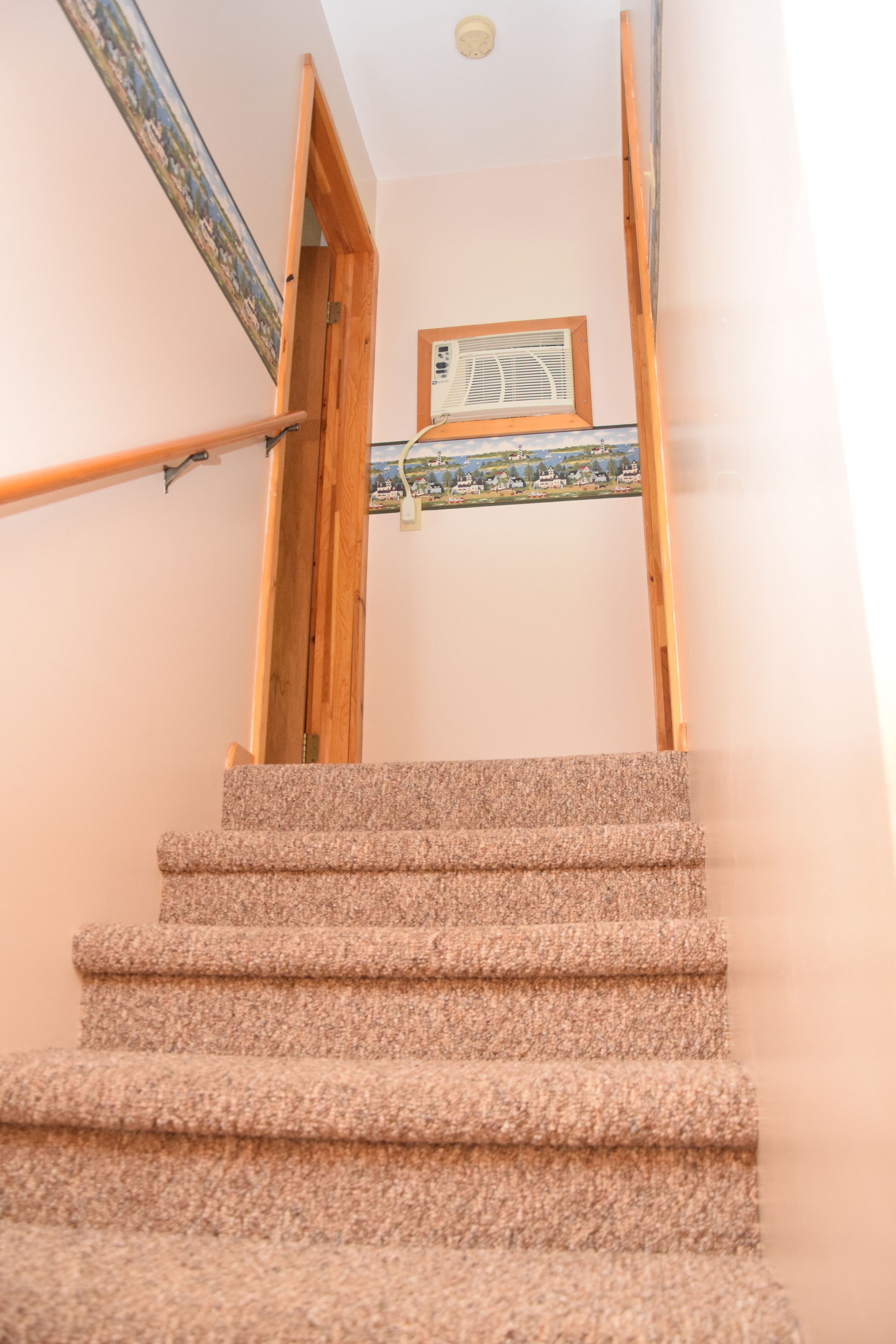 Un escalier recouvert de moquette, doté de rampes en bois et d’une fenêtre offrant une vue décorative sur les maisons.