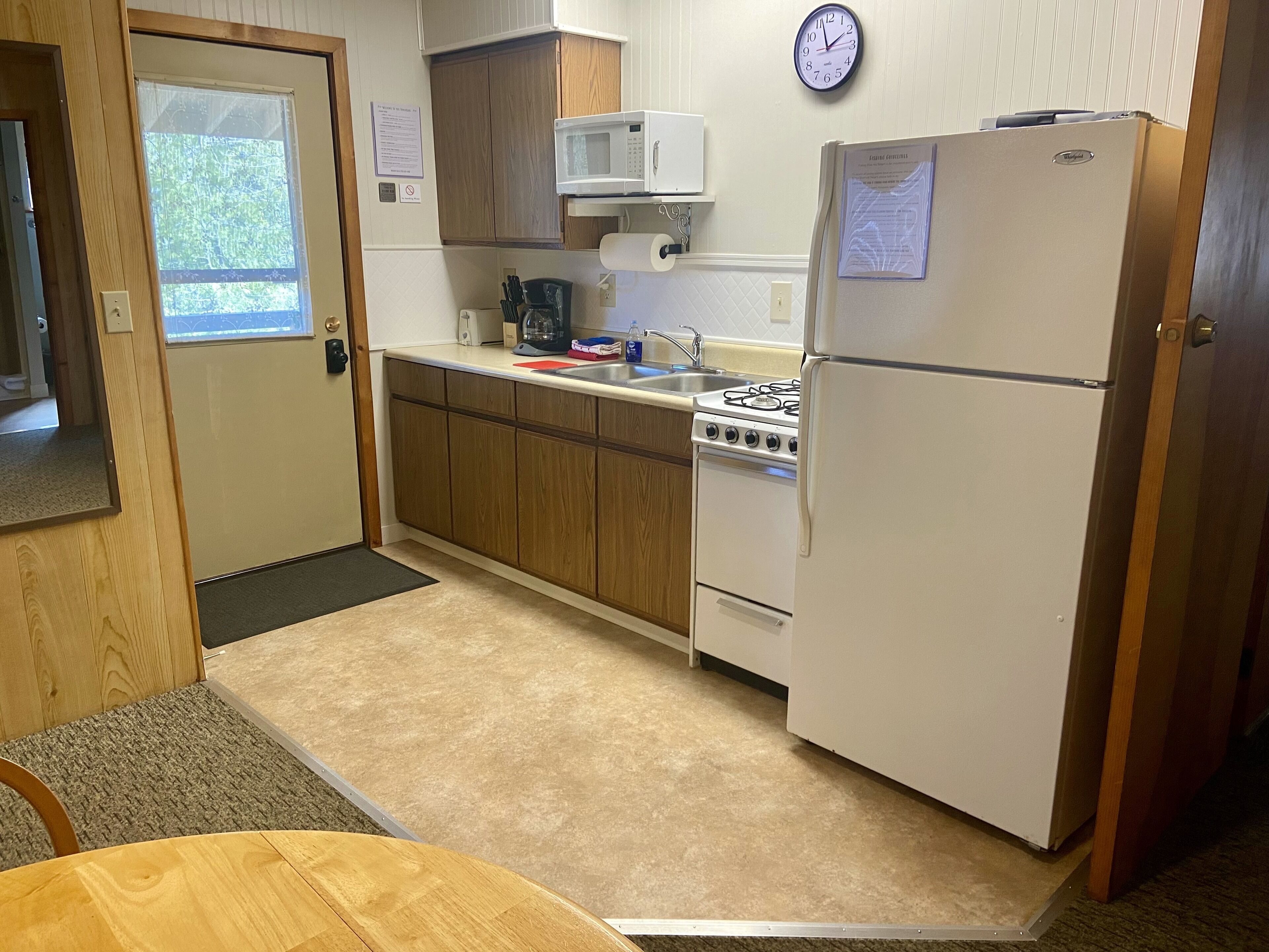 A compact kitchen with wooden cabinets, a white refrigerator, a microwave, a stove, and a clock on the wall.