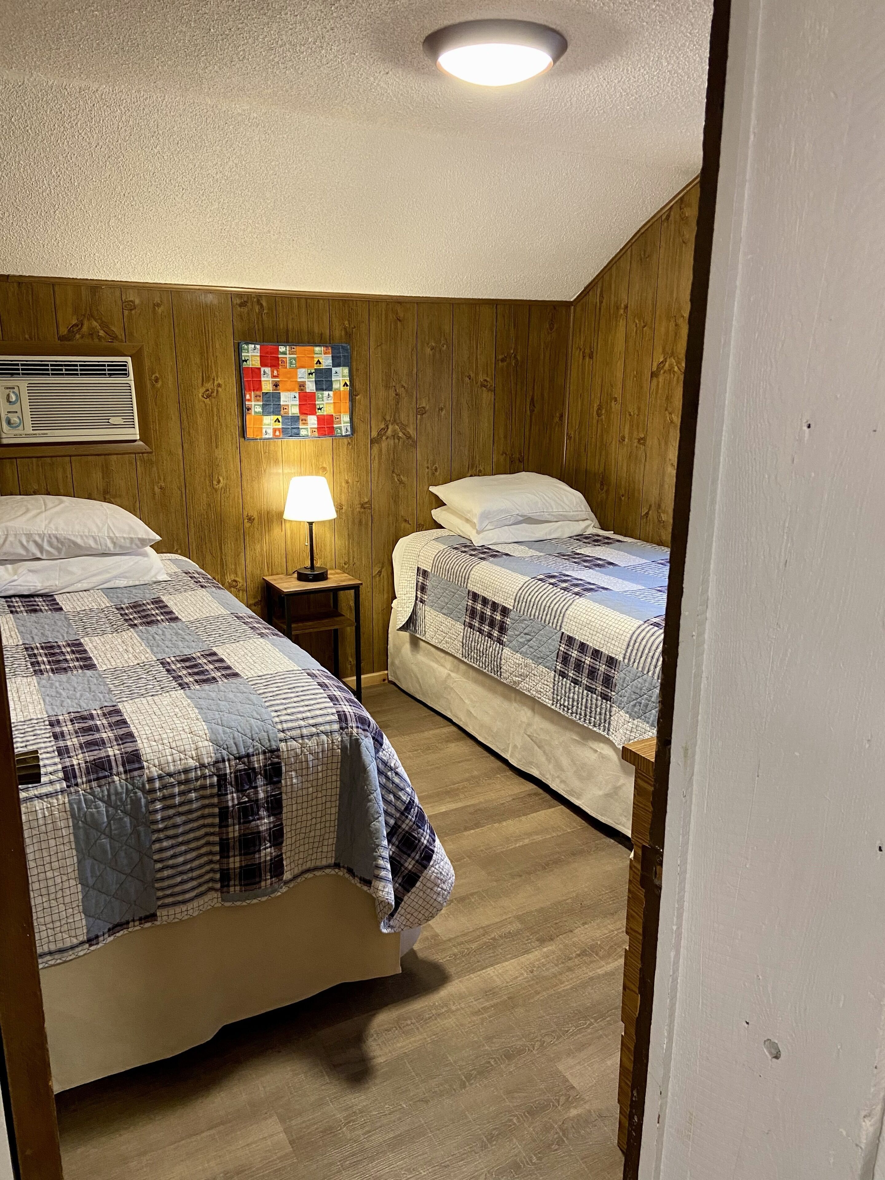 Un dormitorio con dos camas, paredes de madera, colcha acolchada y aire acondicionado montado en la pared, además de un edredón colorido.