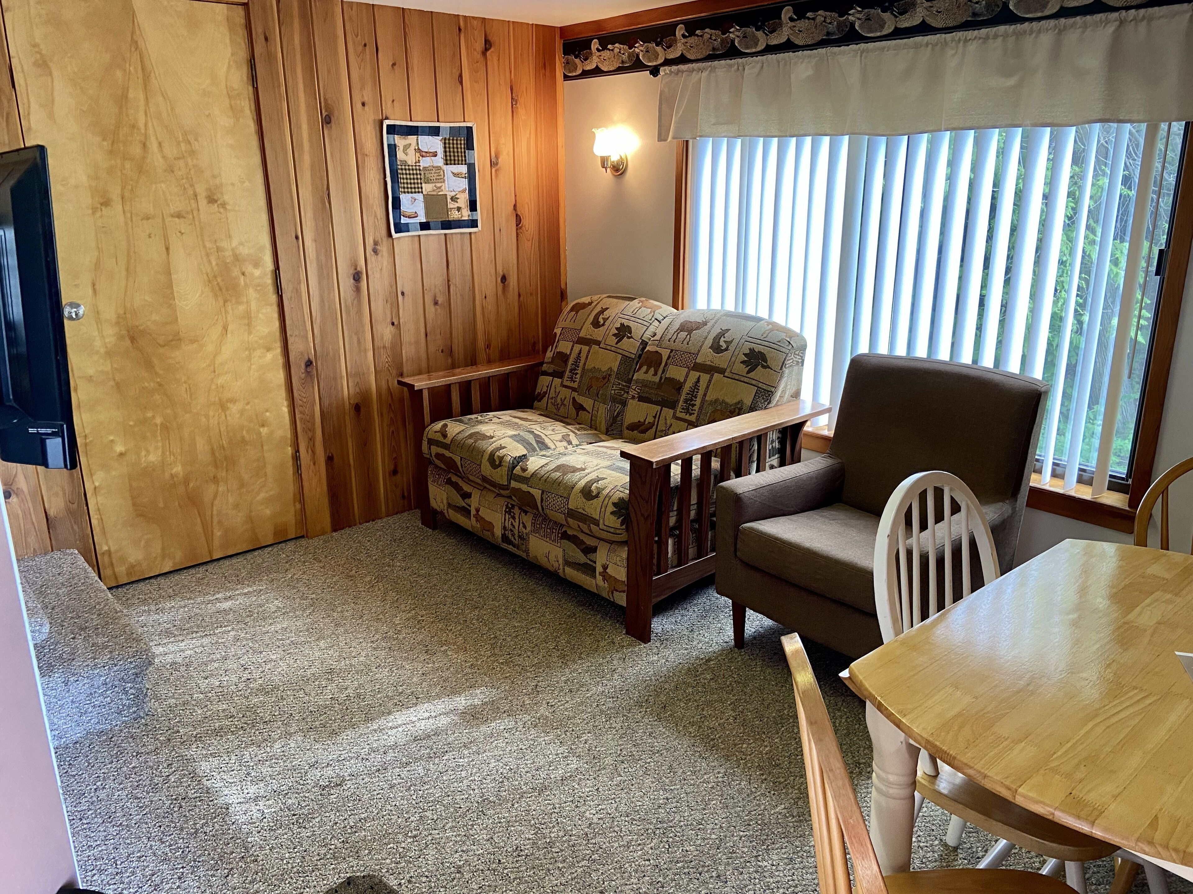 A cozy room with a wooden paneled wall, a patterned sofa, a wooden table, and a window with blinds.