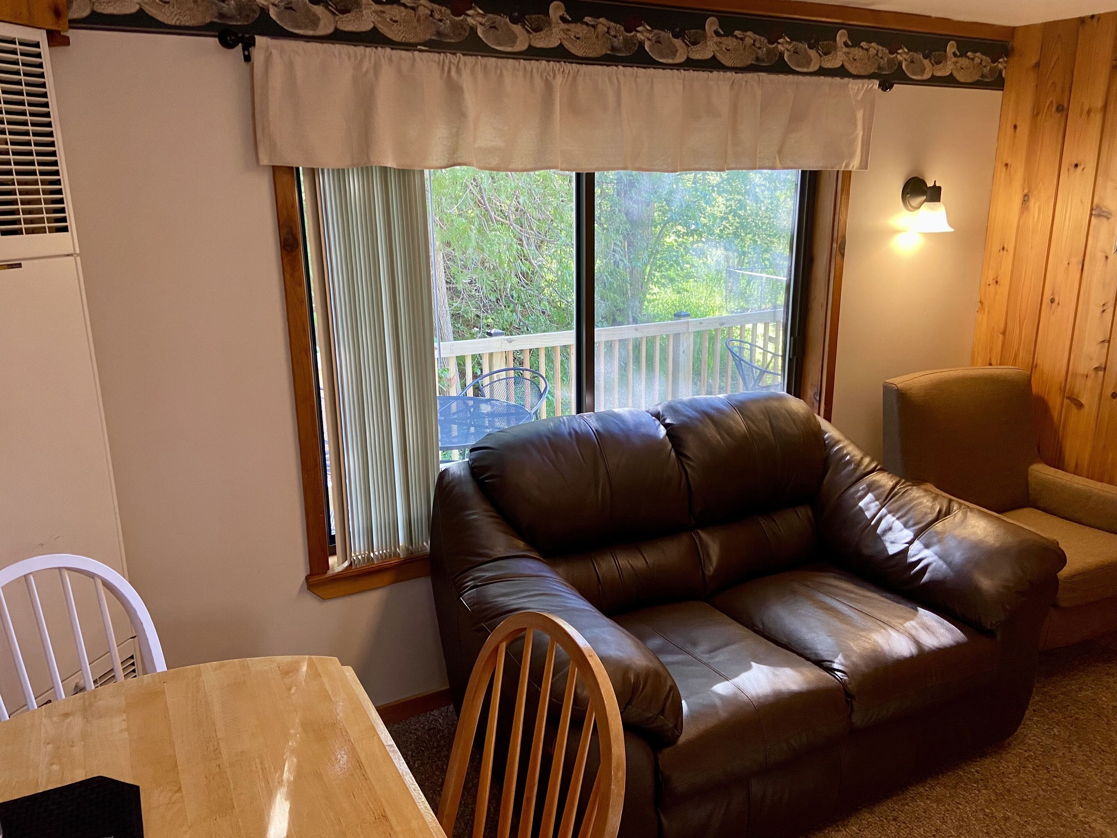 Una habitación con un sofá de cuero marrón, una mesa de comedor de madera, una silla blanca y una ventana con vistas a una terraza de madera y follaje.