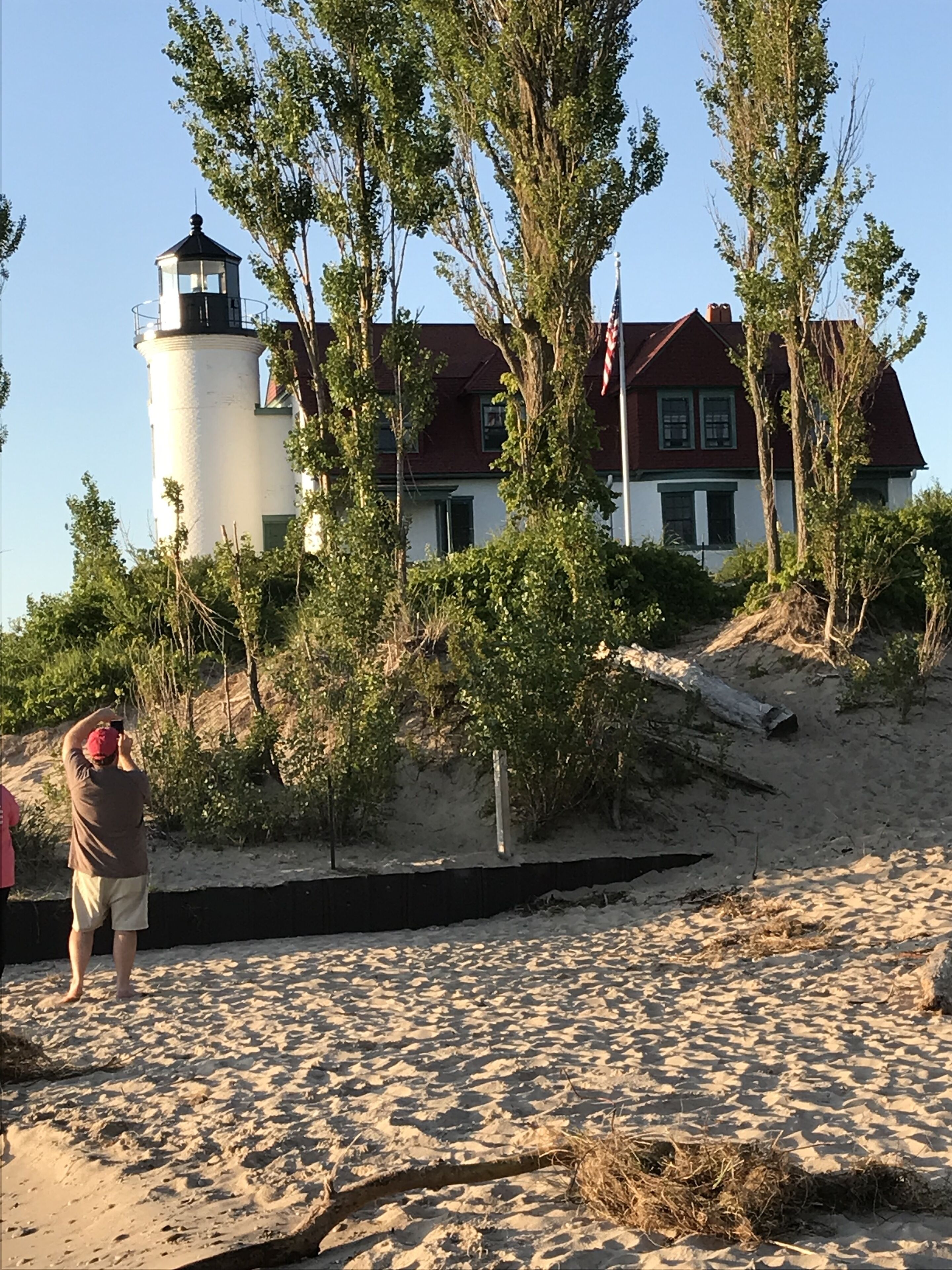 Un phare et une maison avec un toit rouge, entourés d’arbres et de sable.
