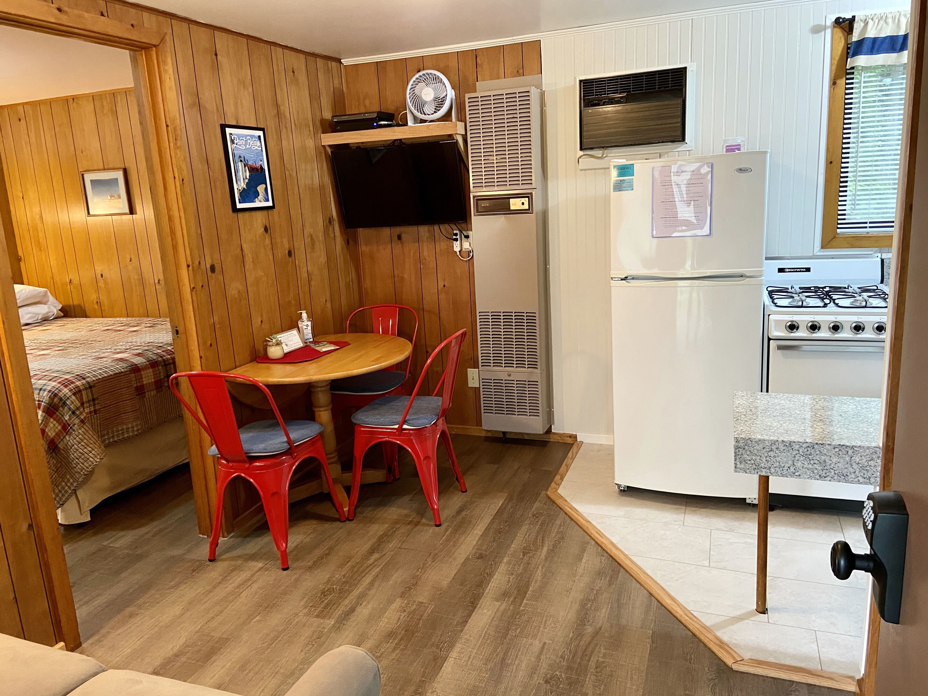 A compact room with a bed, a dining table with red chairs, a small kitchen with a refrigerator, stove, and a wall-mounted fan.