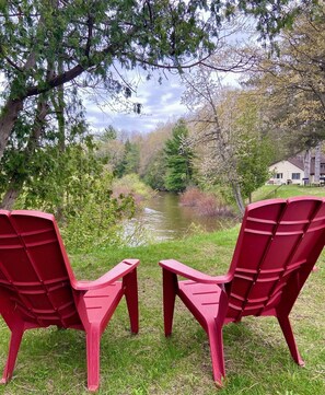 Two red Adirondack chairs facing a pond with trees and a house in the background. - Betsie Riverside Resort (Benzonia)