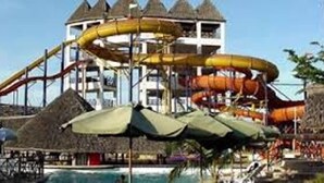 Parque acuático con toboganes y zona de piscina, rodeado de chozas con techo de paja y sombrillas.