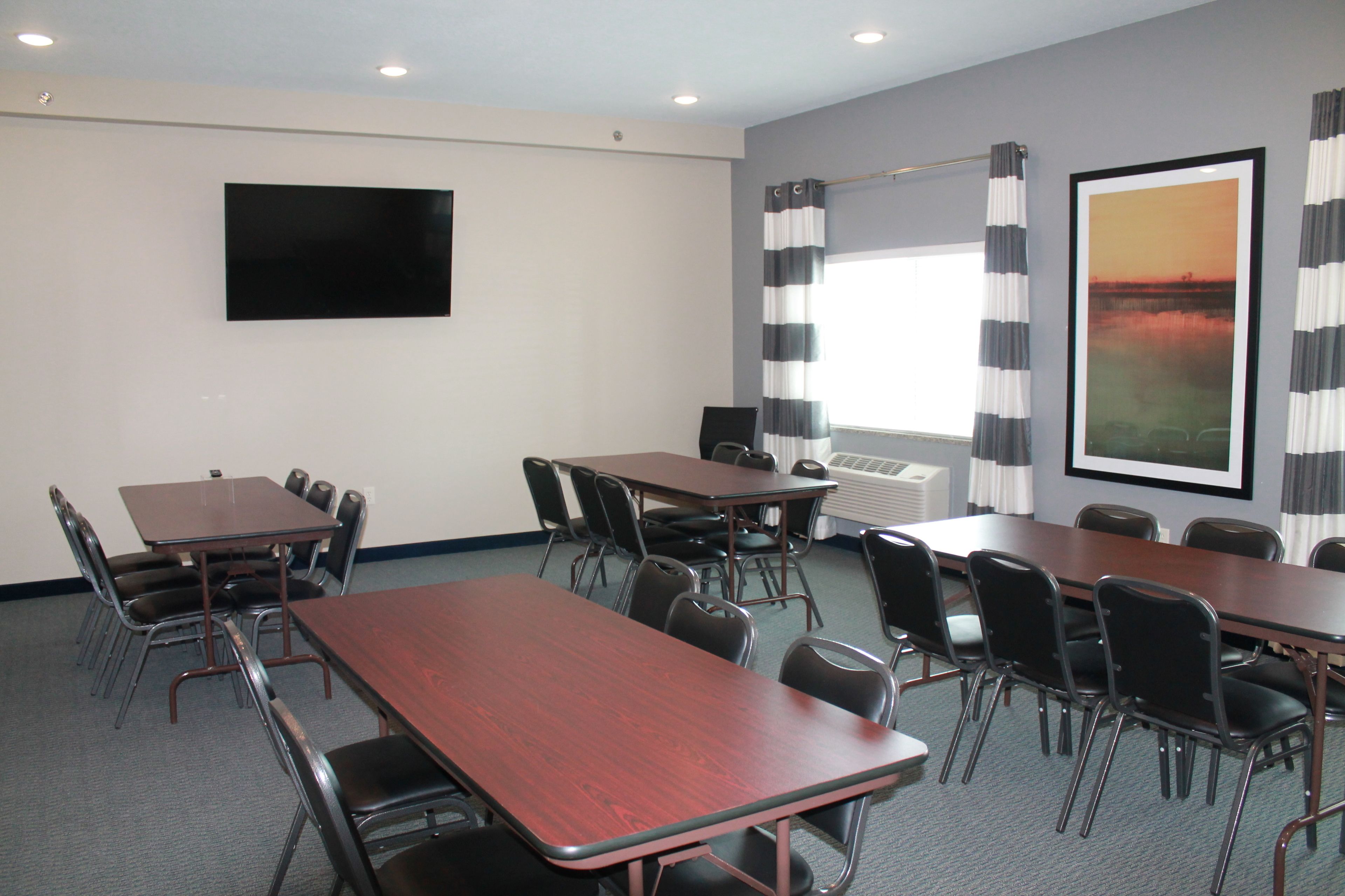 A conference room with a large table, multiple chairs, a flat-screen TV, a window with curtains, and a framed picture on the wall.