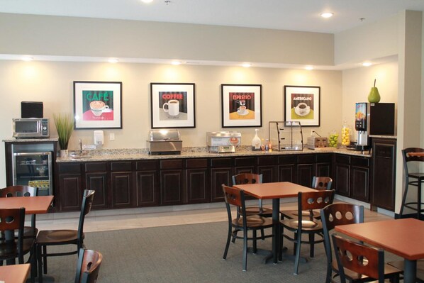 A modern kitchen with dark wooden cabinets, a granite countertop, and a coffee station. There are framed pictures on the wall, a microwave, and a refrigerator. - Reston Lodge & Suites (Emmetsburg)