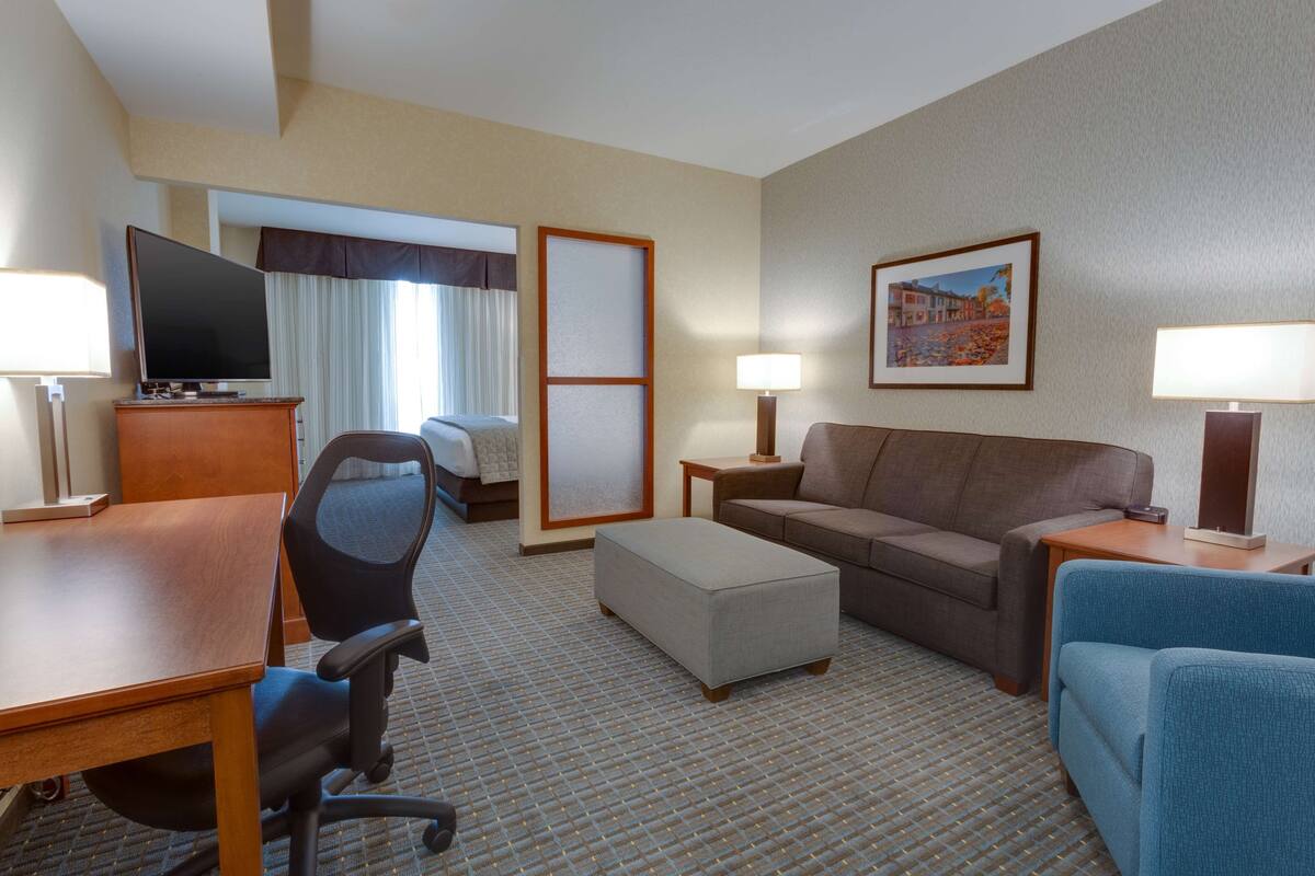 A hotel room with a bed, a desk with a computer, a TV, a sofa, a chair, and a small table with a lamp.