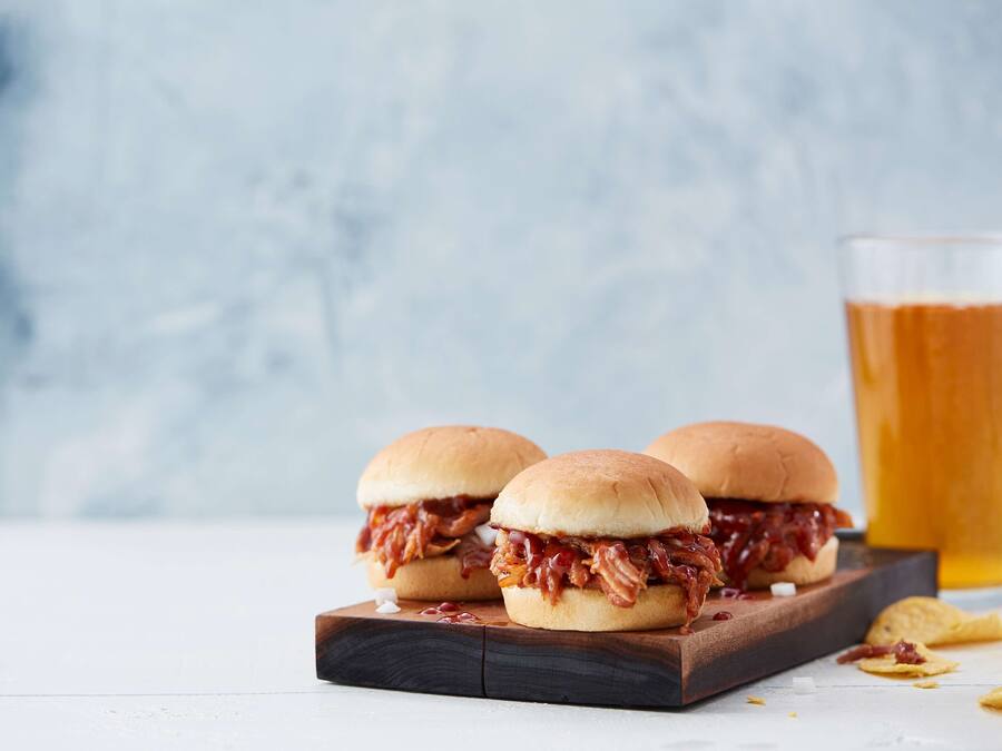 Two pulled pork sandwiches on a wooden board with a glass of beer in the background.