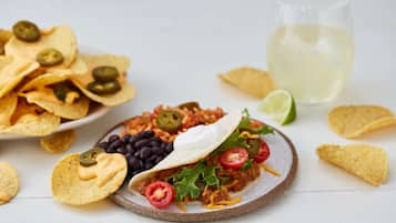 A plate of nachos, a taco with toppings, and a glass of white beverage on a white surface.