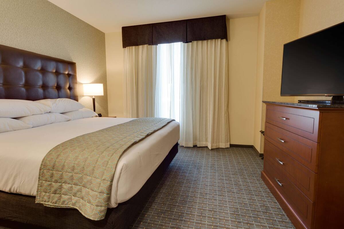 A hotel room with a large bed, a flat-screen TV, a dresser, and a window with curtains.