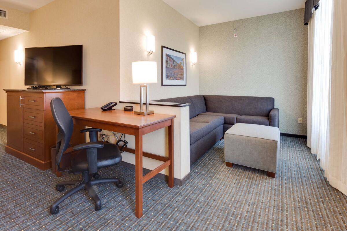 A hotel room with a desk, chair, sofa, ottoman, TV, and a framed picture on the wall.