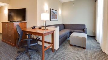 A hotel room with a desk, chair, sofa, ottoman, TV, and a framed picture on the wall.