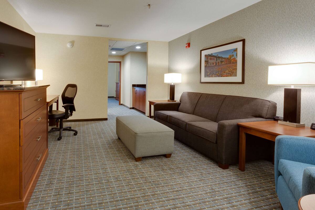 A hotel room with a sofa, ottoman, desk, chair, and a television.