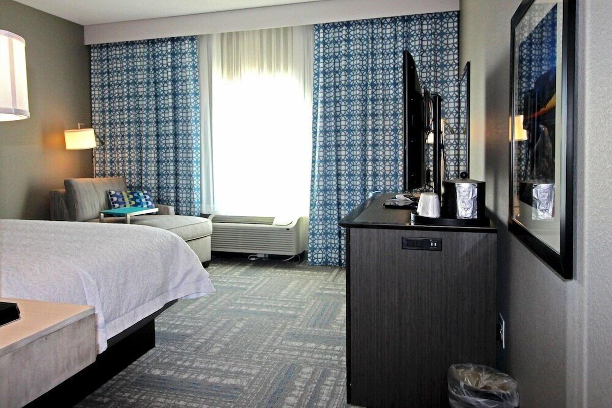 Een hotelkamer met een bed, een bureau, een stoel, een televisie, een minibar en een prullenbak.