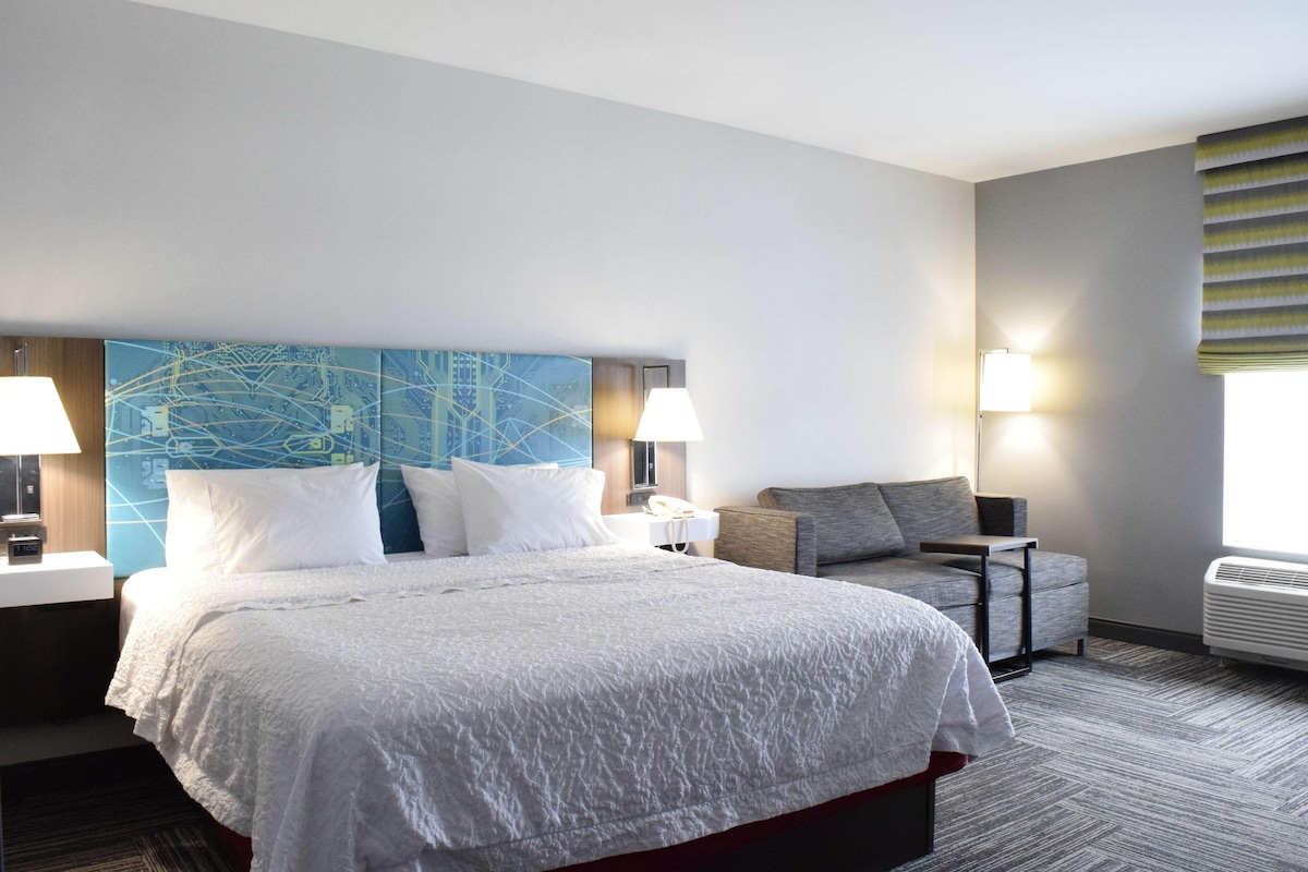 A hotel room with a large bed, a desk, a chair, and a wall-mounted lamp.
