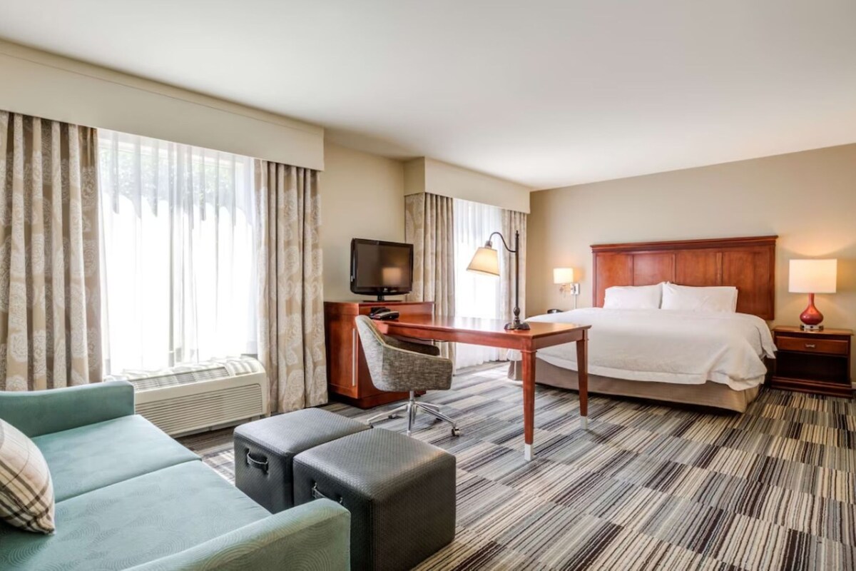 A hotel room with a bed, a desk, a chair, a TV, and a window with curtains.