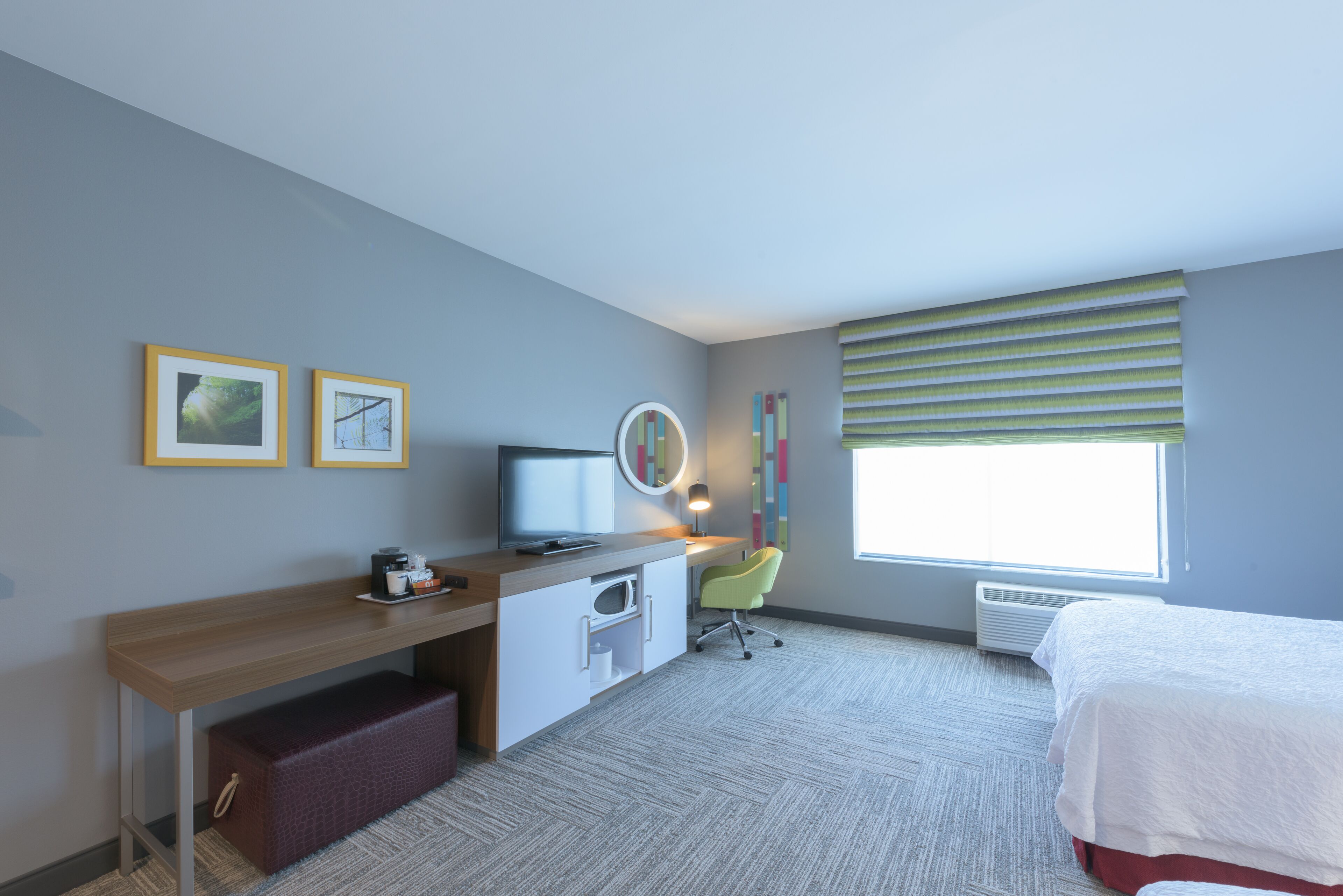 A hotel room with a bed, a desk with a computer, a chair, a small table with a coffee maker, a mirror, and a window with blinds.