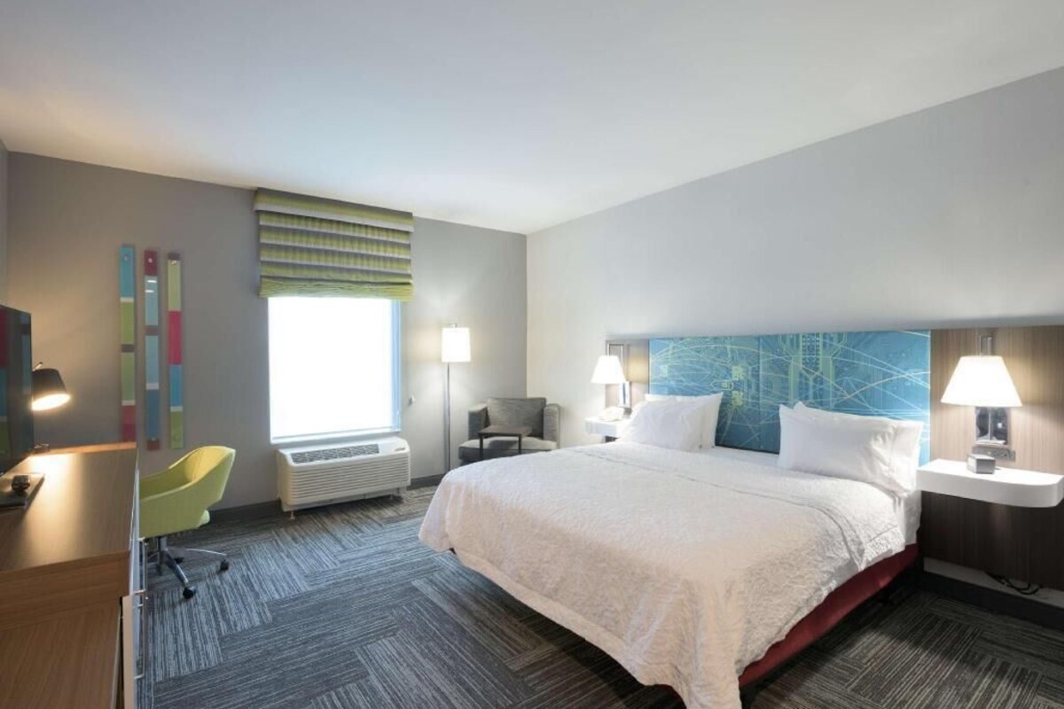 A hotel room with a bed, a desk, a chair, a window with blinds, and a wall with a blue abstract design.
