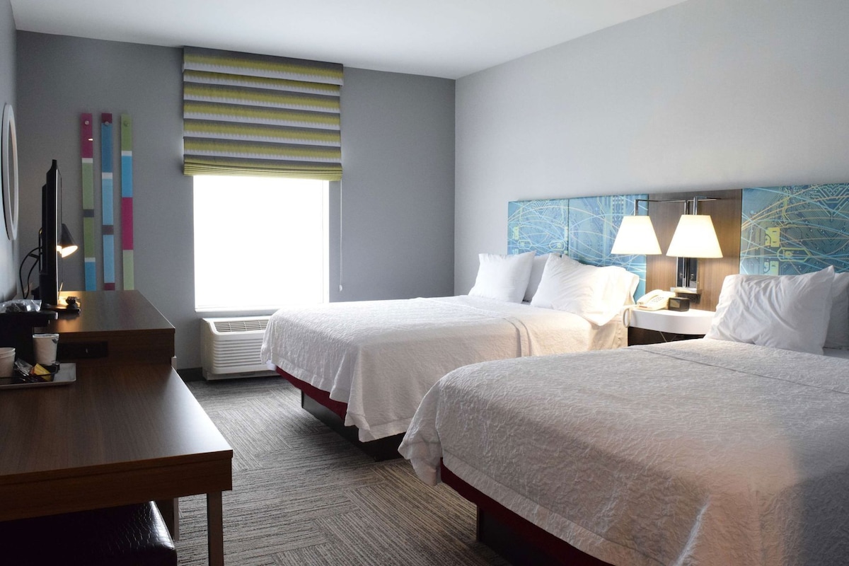 A hotel room with two beds, a desk, a chair, a lamp, a map on the wall, and a window with blinds.