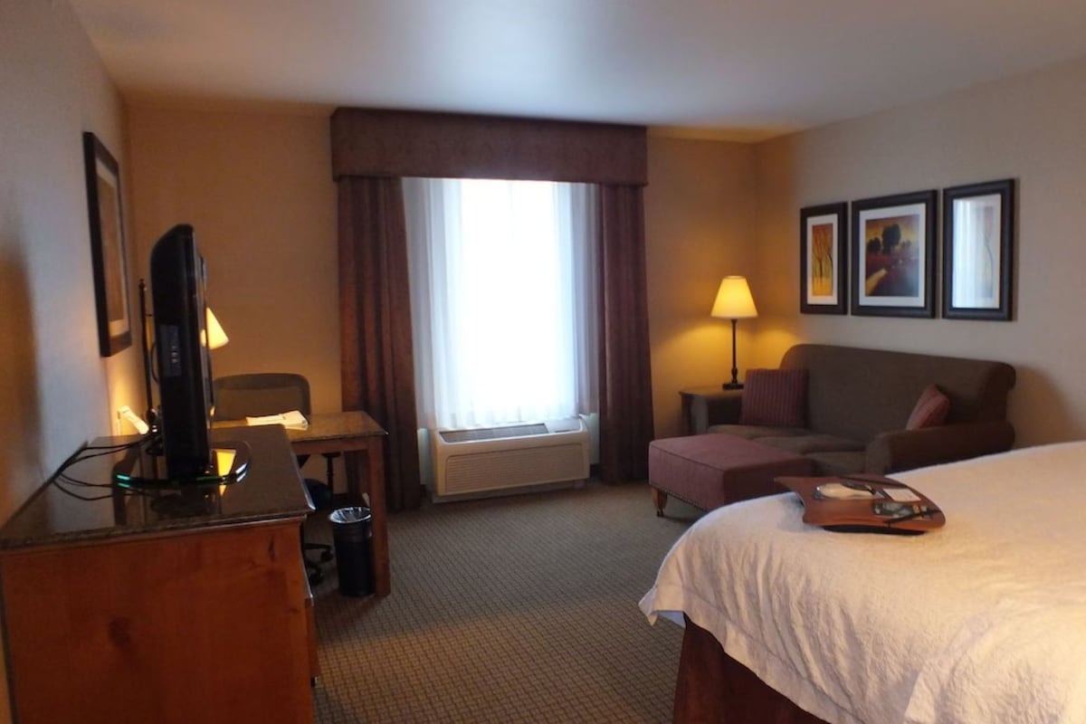 A hotel room with a bed, a desk with a computer, a chair, a lamp, and a window with curtains.