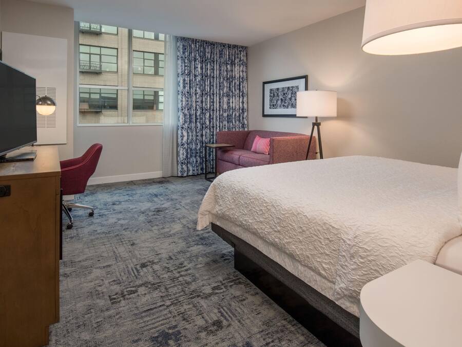 Habitación de hotel con cama, escritorio con televisor, silla, lámpara y ventana con cortinas.