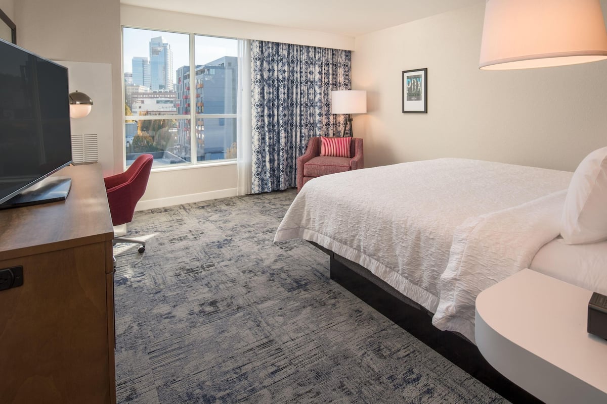 A hotel room with a large bed, a desk with a TV, a red chair, a window with curtains, and a city view.