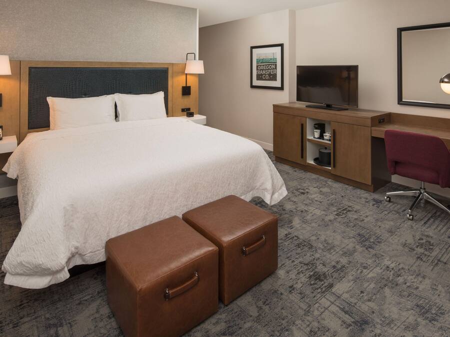 A hotel room with a large bed, a desk with a TV, a chair, and a small table with a lamp.