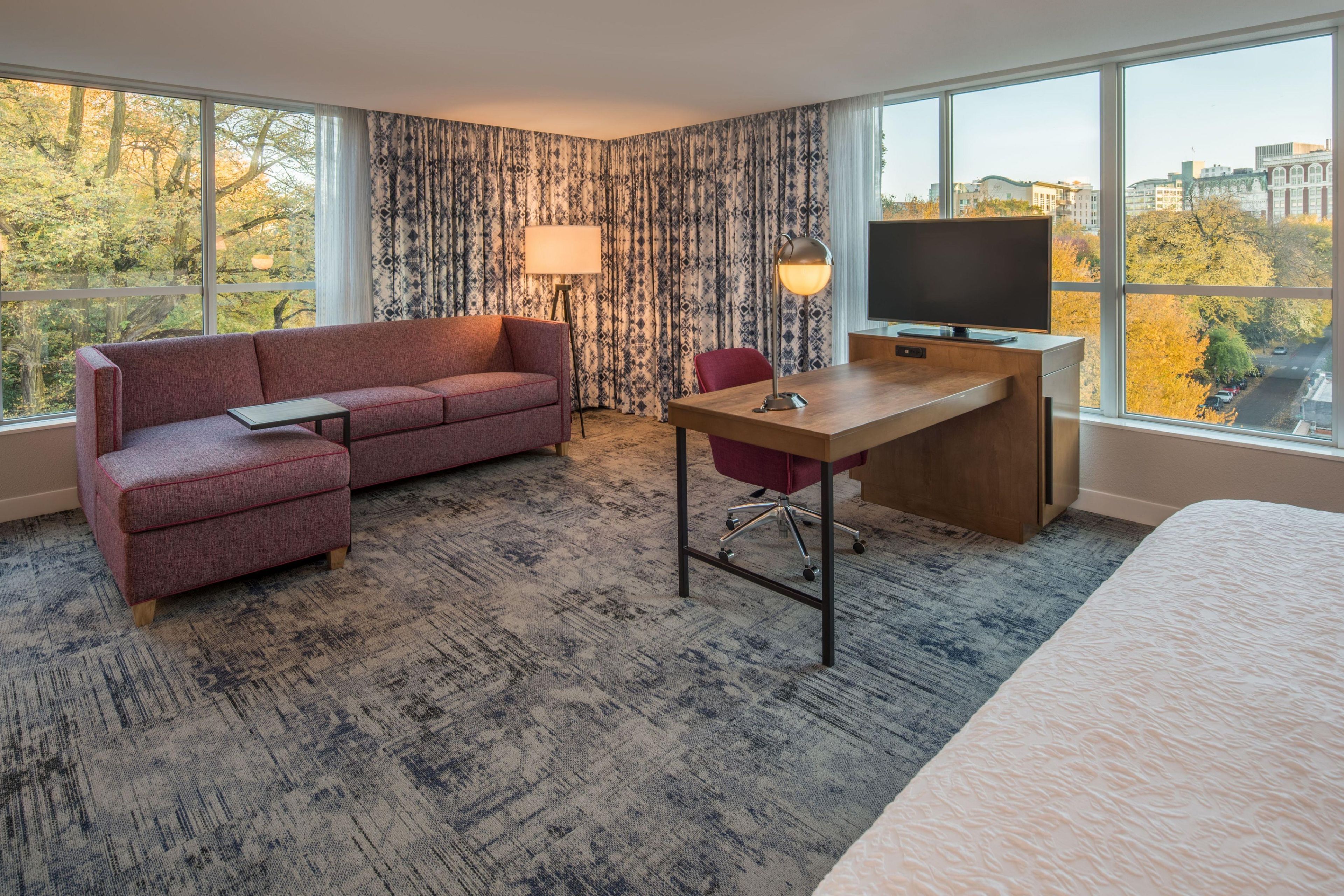 A hotel room with a large window, a sofa, a desk, a chair, and a TV.
