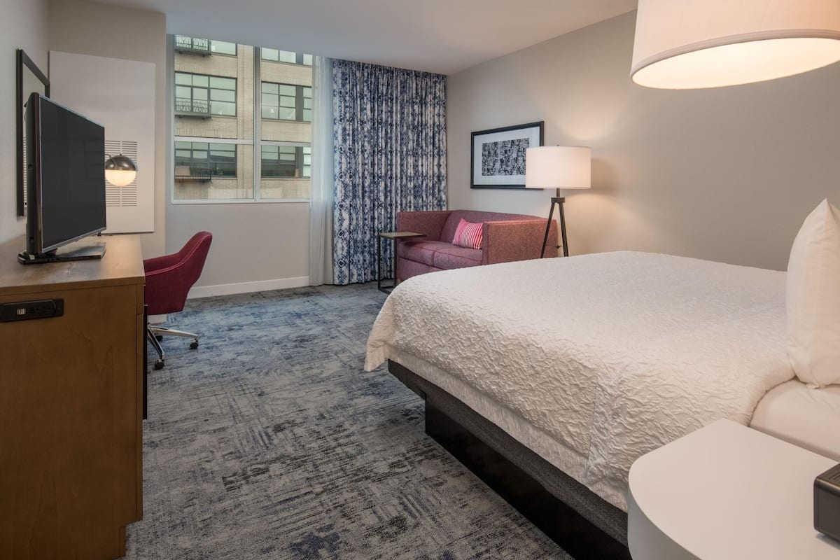 A hotel room with a bed, a desk with a TV, a chair, a lamp, and a window with curtains.