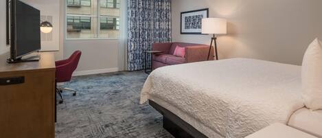 A hotel room with a bed, a desk with a TV, a chair, a lamp, and a window with curtains.