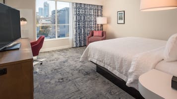 Une chambre d’hôtel avec un grand lit, un bureau avec une télévision, un fauteuil rouge, une fenêtre avec des rideaux et une vue sur la ville.