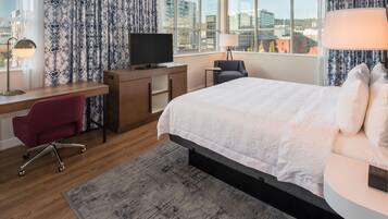 Habitación de hotel con cama, escritorio, televisión, silla y ventanas amplias con vista a la ciudad.
