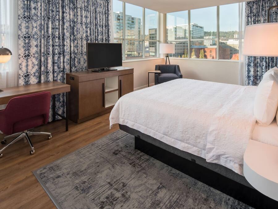 A hotel room with a bed, desk, TV, chair, and large windows with a city view.