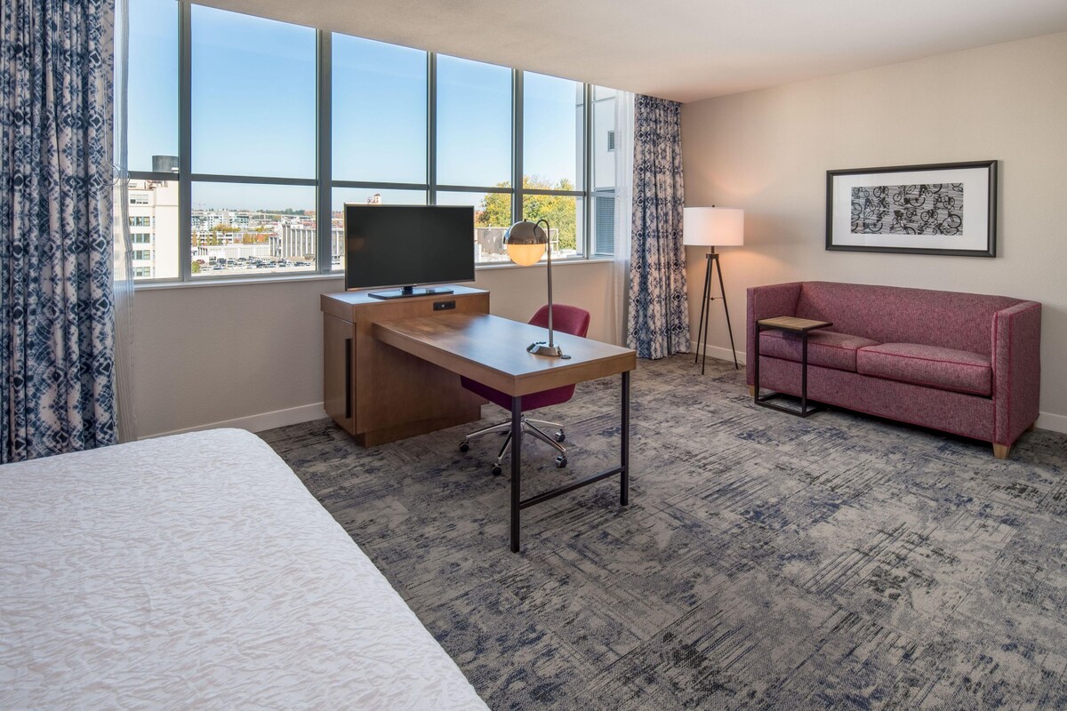 A hotel room with a bed, a desk with a TV, a red chair, a red sofa, a floor lamp, and a framed picture on the wall.
