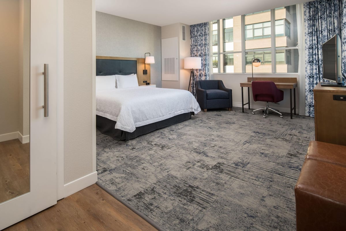 A hotel room with a bed, desk, chair, and large windows.