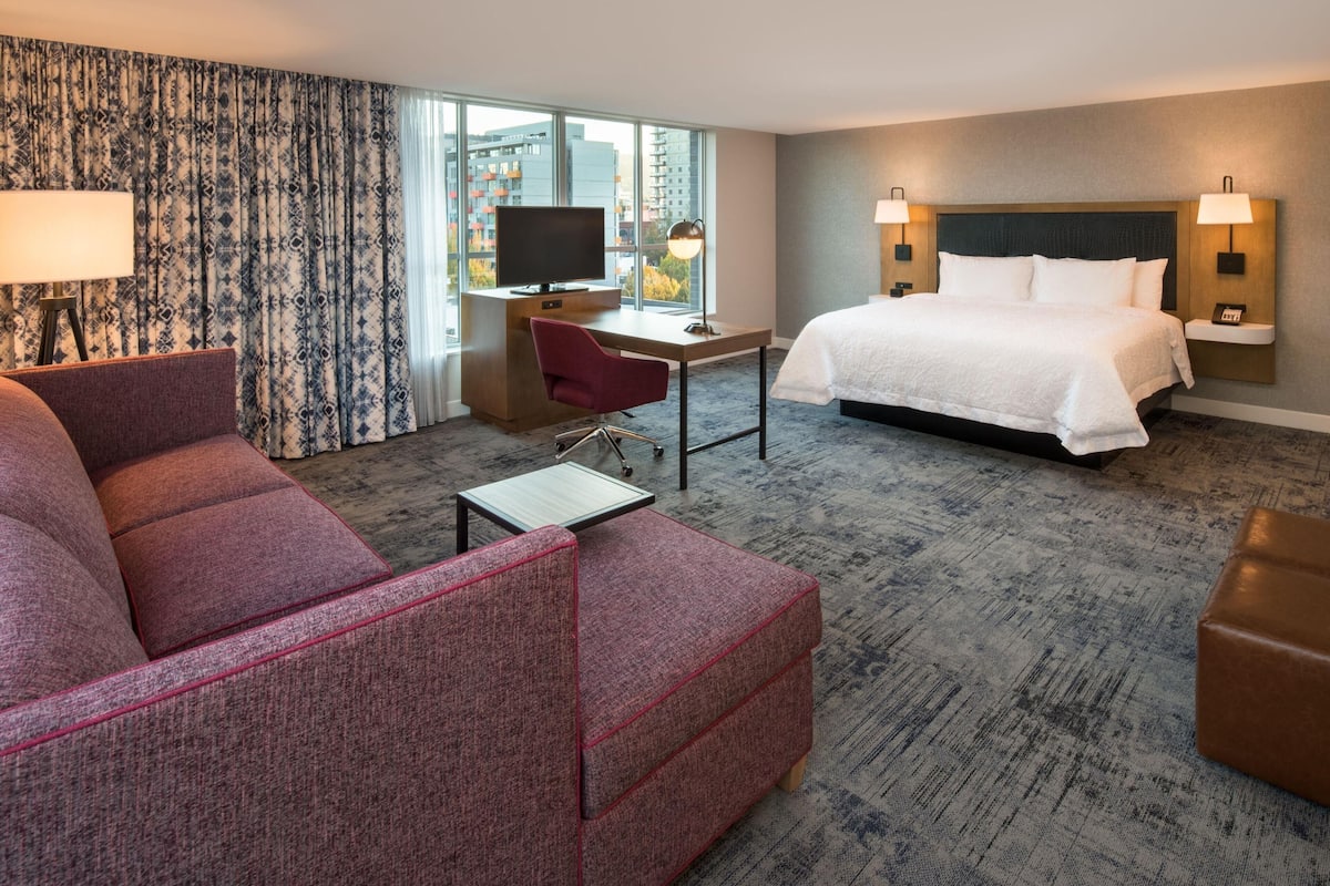 A hotel room with a bed, a desk with a television, a red armchair, and a view of the city through large windows.