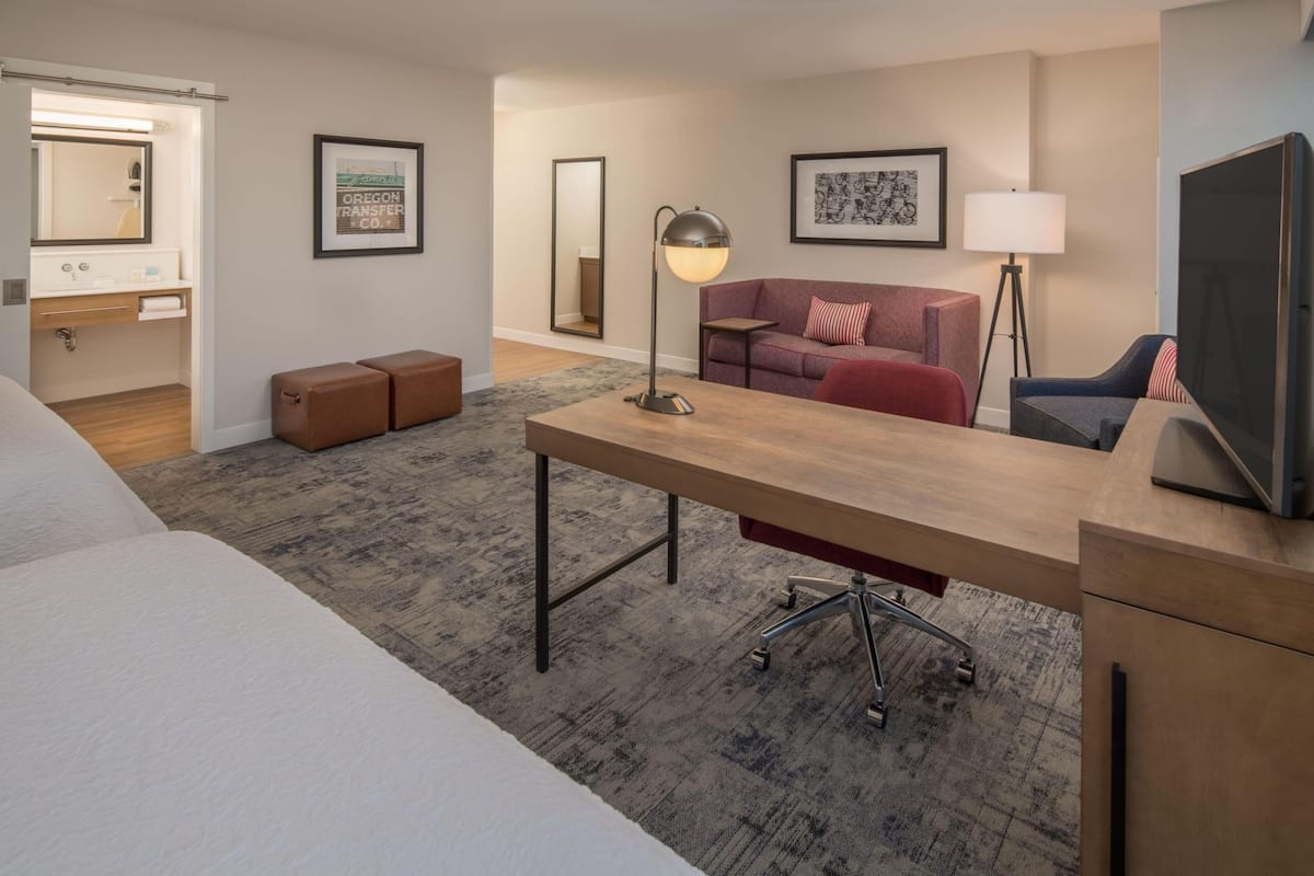 A hotel room with a bed, a desk, a sofa, a TV, a lamp, a mirror, and a framed picture on the wall.