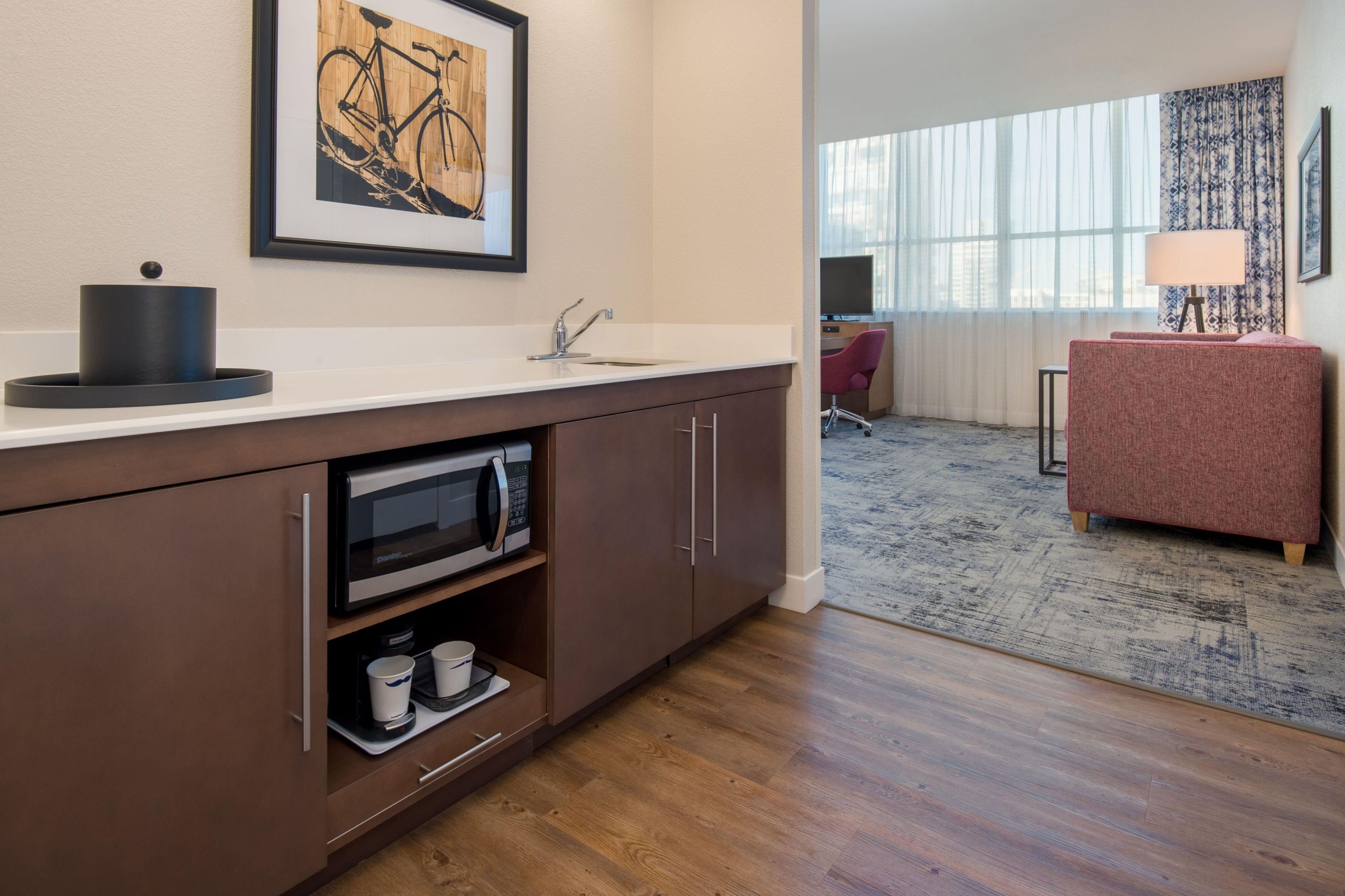 A modern kitchen with a microwave, sink, and cabinets. There is a framed artwork on the wall, a rug on the floor, and a desk with a lamp.