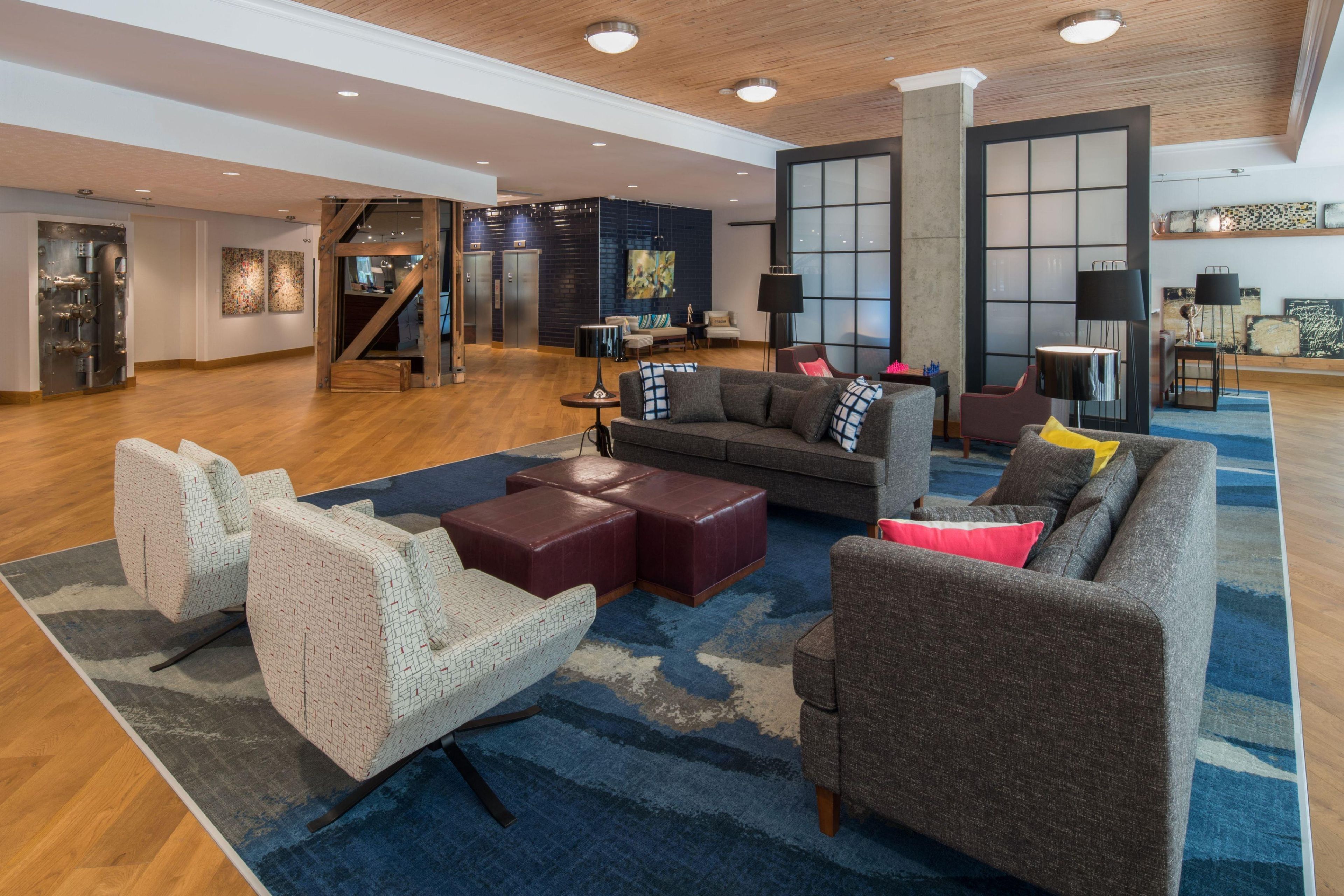 A spacious lobby with a modern design, featuring a large sofa, armchairs, and a coffee table. There are two elevators and a staircase leading to another floor.