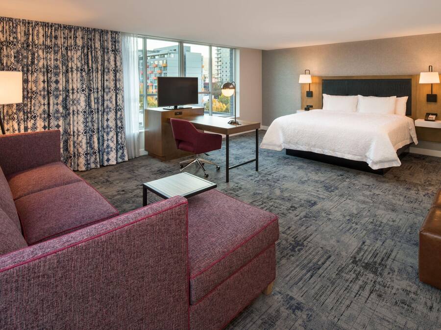 A hotel room with a bed, a desk with a television, a red armchair, and a view of the city through large windows.