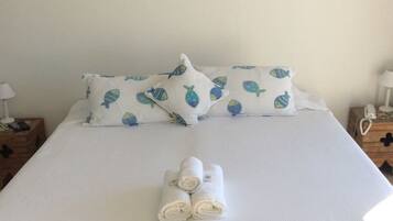 Una cama bien hecha con cubrecama y almohadas blancas a juego, un cuadro en la pared y una mesita de noche de madera con una lámpara.