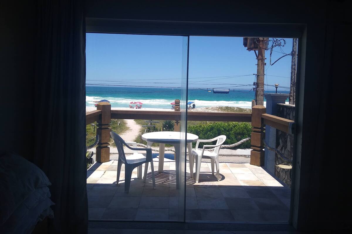 Un balcón con mesa y sillas, con vista a una playa con gente y sombrillas.