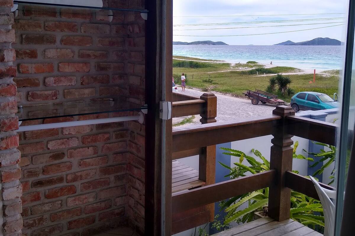 Una habitación con pared de ladrillo, puerta de cristal que da a un balcón con barandilla de madera, y vista al mar y la playa.