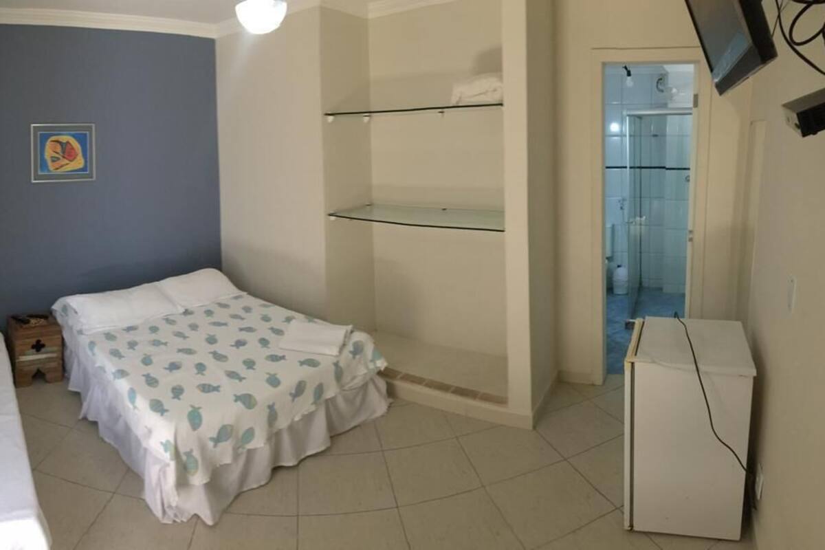 Una habitación pequeña y funcional con cama, mesita de noche, televisor montado en la pared y puerta de acceso al baño.