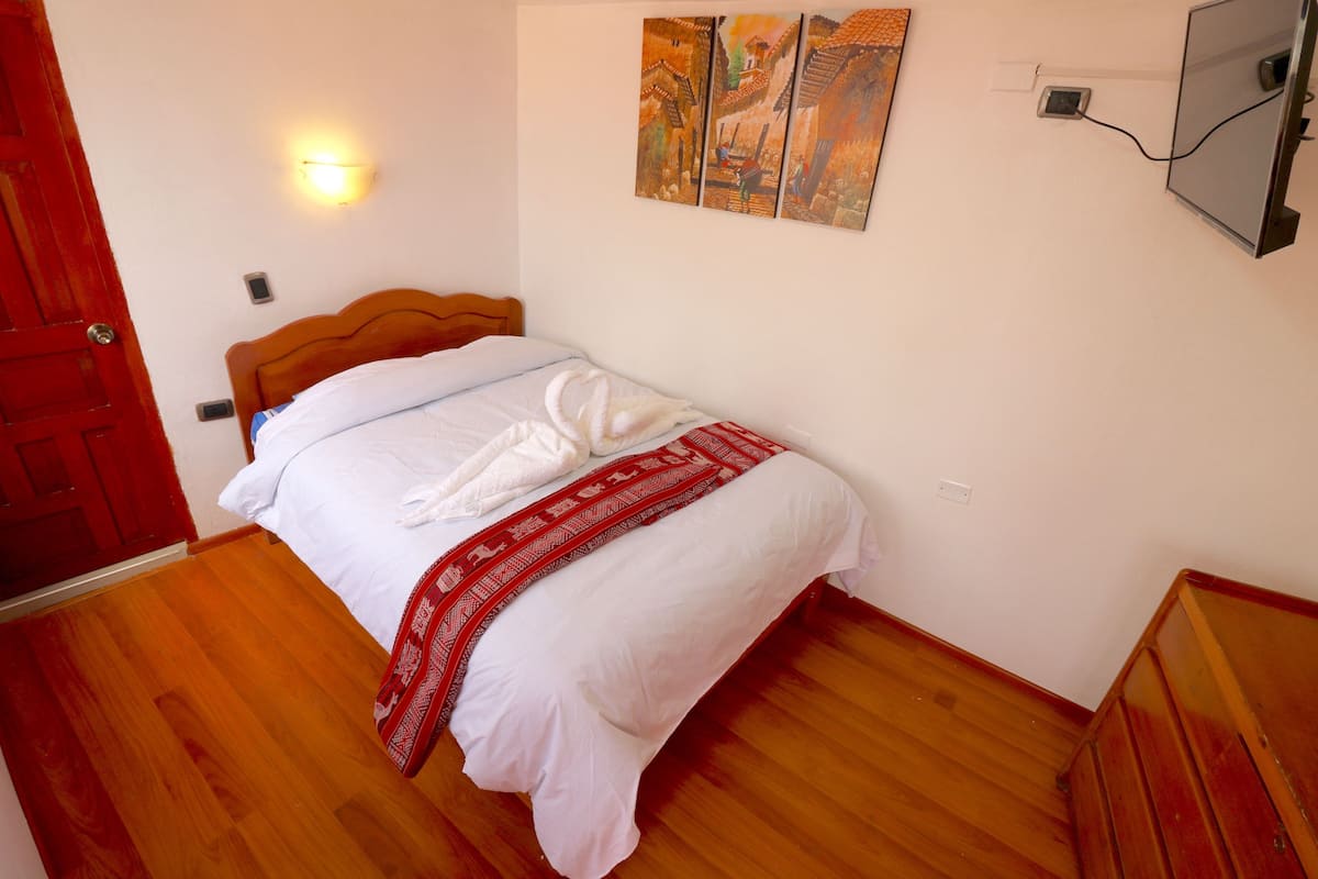 A bedroom with a wooden floor, a bed with white bedding and a red blanket, a wooden headboard, a wall-mounted light, and a painting above the bed.