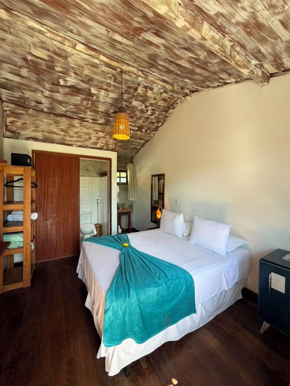 Ein Schlafzimmer mit Holzboden, einem Bett mit weißer Bettwäsche, einem Schrank aus Holz und einem durch eine offene Tür einsehbaren Badezimmer.
